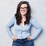 Promotional image of woman standing in front grey wall smiling with her hands on her hips