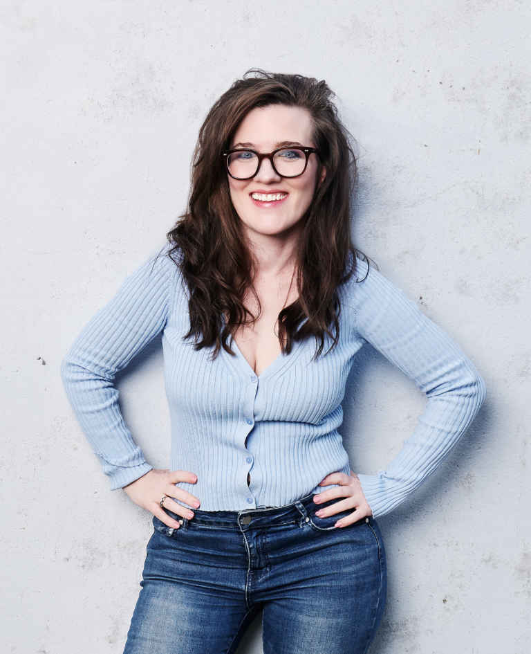 Promotional image of woman standing in front grey wall smiling with her hands on her hips